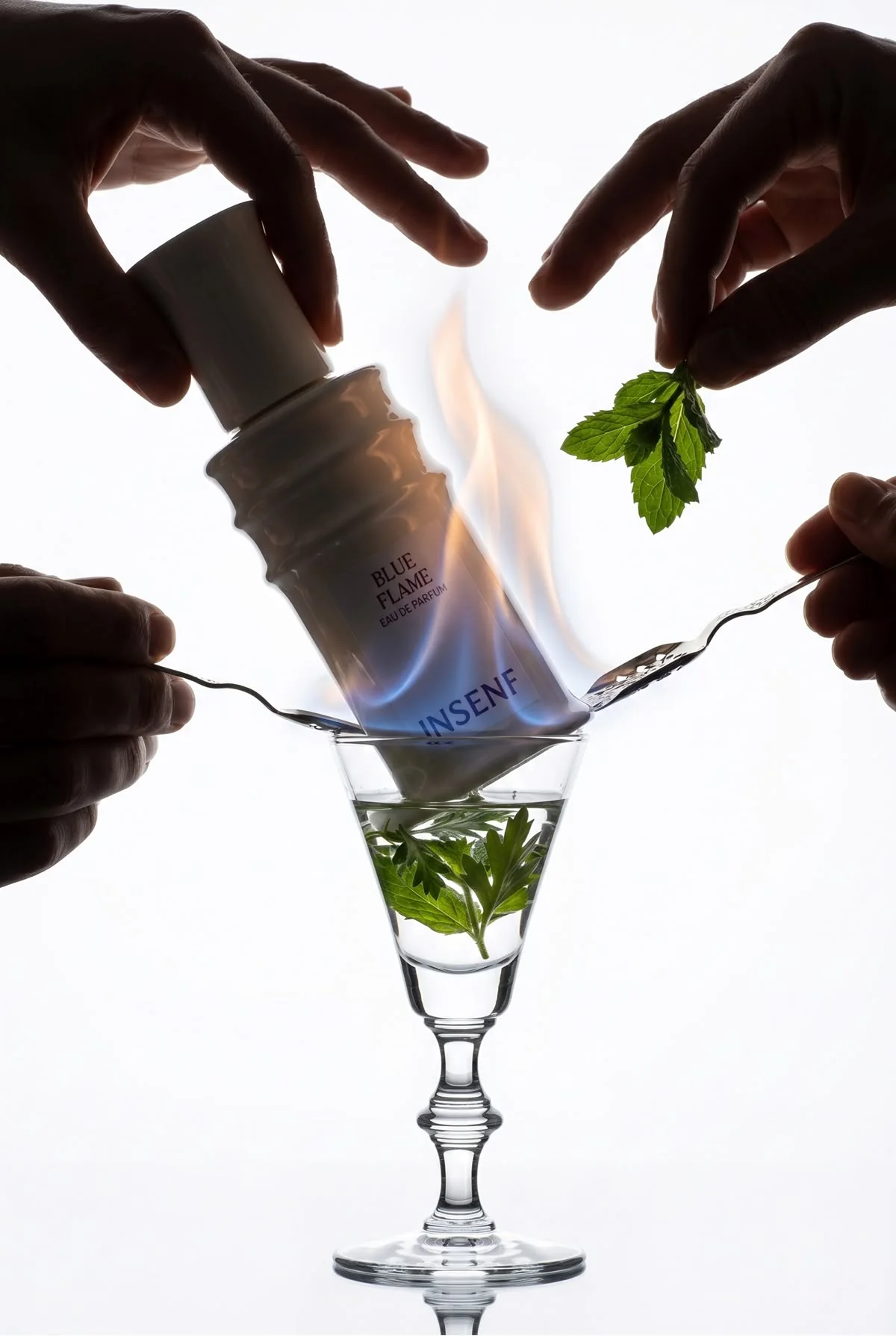 Mains tenant le flacon de Blue Flame renversé au-dessus d'un petit verre à pied, dans une flamme bleue, avec cuillères et menthe